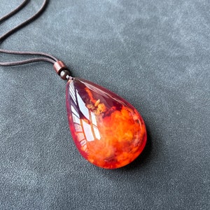 May include: A teardrop-shaped pendant necklace with a brown cord. The pendant is made of amber with a reddish-orange hue and contains inclusions of plant matter.