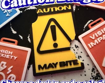 May include: A rectangular yellow and black caution badge featuring a black triangle with an exclamation point and the text "AUTION" at the top and "MAY BITE" at the bottom. The badge has a notched edge and is surrounded by other similar rectangular badges.