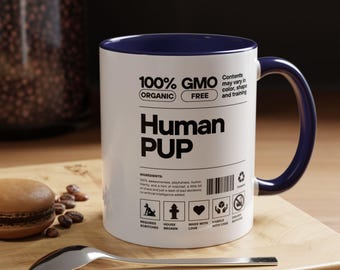 May include: A white ceramic mug with a dark blue interior and handle, featuring a black nutritional label graphic that reads 100% GMO FREE, Human PUP, and lists ingredients like playfulness and humor, along with various icons and text.