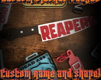 May include: A white and black knife-shaped badge featuring the red text REAPER. The badge has a black handle with two white circular accents. The design is displayed alongside other badge accessories on a dark wooden surface.