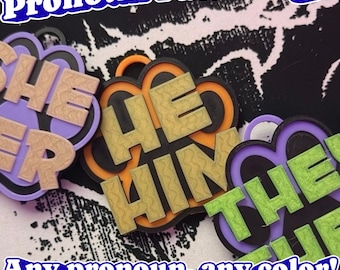 May include: Three paw-shaped badges with raised lettering. One badge is purple with tan 'SHE HER' text, one is orange and black with gold 'HE HIM' text, and one is purple and black with green 'THEY THEM' text. Text reads 'Pronoun Paw Badge'.