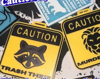 May include: A yellow and black square plastic badge featuring a graphic of a raccoon face above the text TRASH THEIF. The badge has a top loop for attachment. Another badge shows a lion graphic with the text CAUTION and MURDER.