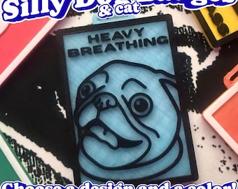 May include: A rectangular badge featuring a light blue 3D-printed pug face with black outlines and the text HEAVY BREATHING in black. The badge has a black border and a small loop at the top for attachment, showcasing a detailed graphic design.