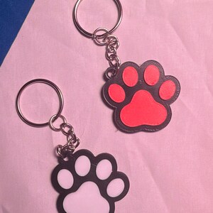 May include: Two keychains featuring a paw print design. One keychain has a black border with a bright pink center, and the other has a black border with a white center. Each keychain includes a silver-colored metal split ring and a short chain.