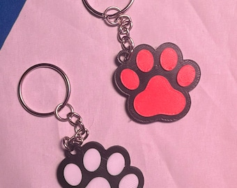 May include: Two keychains featuring a paw print design. One keychain has a black border with a bright pink center, and the other has a black border with a white center. Each keychain includes a silver-colored metal split ring and a short chain.