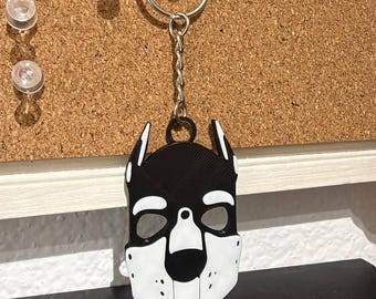 May include: A black and white keychain featuring a stylized dog mask design. The pendant is suspended from a silver-toned metal split ring by a short chain. The mask has pointed ears, eye cutouts, and a distinct snout section with small dots.
