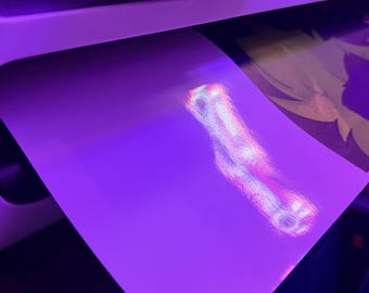 May include: A white printer tray holding a sheet of glossy paper with a bright, iridescent light reflection in the center. To the right, a partially visible printed graphic features green and dark purple shapes under purple ambient lighting.
