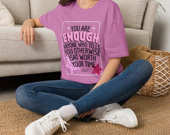 May include: A light purple t-shirt featuring a white rectangular border containing the black and pink text: YOU ARE ENOUGH ANYONE WHO TELLS YOU OTHERWISE ISNT WORTH YOUR TIME -DOLLY, accented by two glittery pink star graphics on the lower corners.
