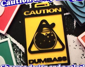 May include: A rectangular yellow and black badge featuring the text CAUTION at the top and DUMBASS at the bottom. In the center, a black triangle contains a graphic of a cat inside a fishbowl. The badge has a small loop at the top for attachment.