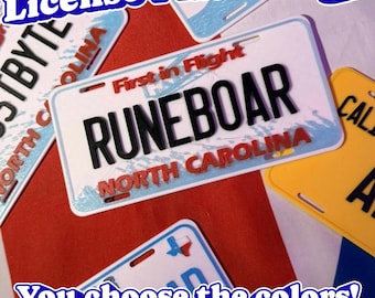 May include: A rectangular North Carolina license plate badge featuring the text RUNEBOAR in black, with the red text First in Flight and North Carolina printed on a white background with blue accents and mounting slots at the top and bottom edges.