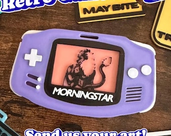 May include: A purple handheld game console badge featuring a white directional pad and two white buttons. The screen displays a black graphic of a creature with a tail and the text MORNINGSTAR in white letters on a black background.