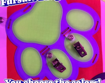 May include: A purple paw-shaped badge holder with light green inserts and two metal clips attached. The text "Fursuit Badge Holders" appears at the top, and "You choose the colors! We print them to order!" is printed at the bottom of the image.