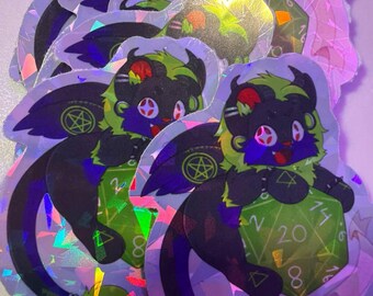 May include: A stack of holographic stickers featuring a black creature with green fur accents, wings, and a long tail. The creature is holding a large green twenty-sided die with visible numbers and a pentagram symbol on its wing.