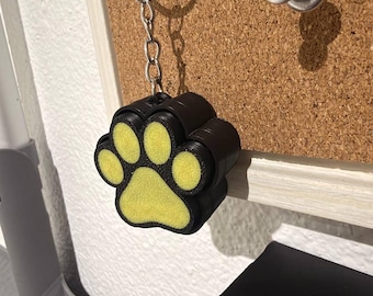 May include: A black keychain featuring a paw print design with a textured yellow center. The paw print is attached to a silver metal split ring by a short silver chain. The object has a thick, layered construction with a matte black finish.