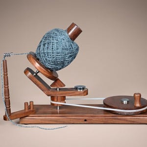 Peut inclure: Un dévidoir à laine en bois foncé composé d'une base horizontale, d'un disque circulaire à manivelle relié par une courroie blanche à une broche rotative et d'un bras de guidage vertical. Une grosse pelote de laine bleu-gris est sur la broche.