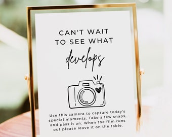May include: A gold-framed sign with the text "CAN'T WAIT TO SEE WHAT develops" and a black and white illustration of a camera with a heart on it. The sign also includes instructions for using the camera and leaving it on the table when the film runs out.