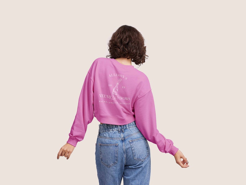 May include: A pink cropped sweatshirt with long sleeves and a white graphic on the back that says "Allison's Velvet Vows Bachelorette Weekend 20 21".