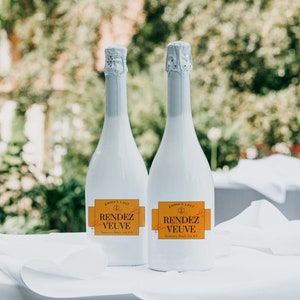 May include: Two white bottles of sparkling wine with orange labels that read "Emma's Last Rendez Vous Veuve" on a white tablecloth.