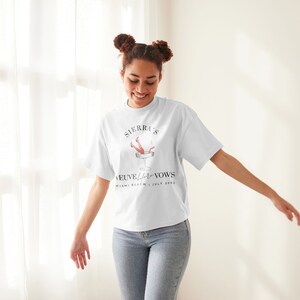 May include: A white t-shirt with a graphic of a woman's legs sticking out of a champagne glass. The text on the shirt reads "Sierra & [Name] Veuve Before Vows Miami Beach | July 2023".