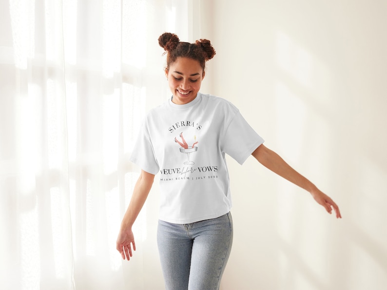 May include: A white t-shirt with a graphic of a woman's legs sticking out of a champagne glass. The text on the shirt reads "Sierra & [Name] Veuve Before Vows Miami Beach | July 2023".