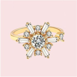 Può includere: Anello dorato con una gemma centrale rotonda e trasparente, circondata da gemme trasparenti più piccole e pietre taglio baguette. L'anello ha un design a raggiera ed è posto su sfondo rosa.