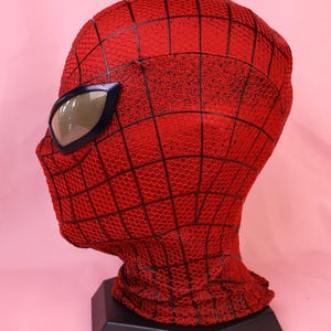 May include: A red full-head mask featuring a black web pattern across the surface. The mask includes large, angular eye lenses with a metallic gold mesh finish and a dark frame, displayed on a black hexagonal stand for a clear profile view.