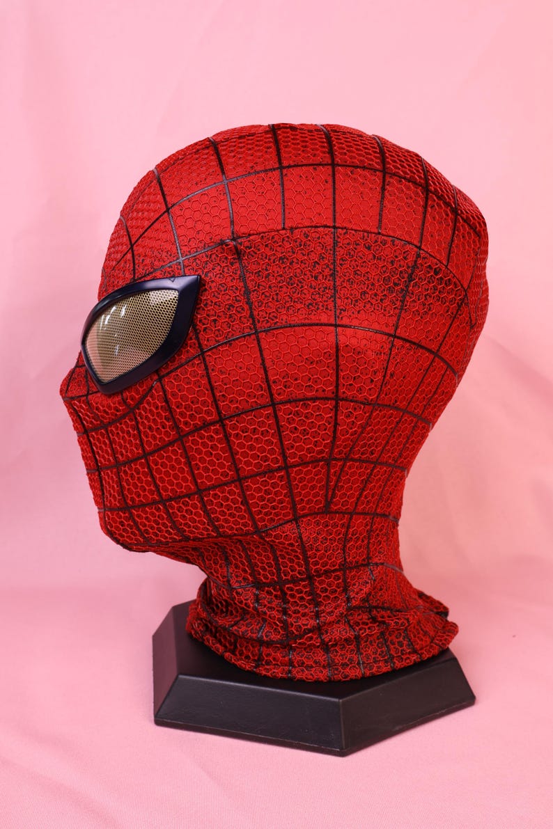 May include: A red full-head mask featuring a black web pattern across the surface. The mask includes large, angular eye lenses with a metallic gold mesh finish and a dark frame, displayed on a black hexagonal stand for a clear profile view.