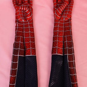 May include: A pair of long, elbow-length gloves featuring a red fabric base with a black spiderweb pattern printed in silver lines. The lower section of each glove is solid black with a textured, grid-like surface pattern throughout the material.