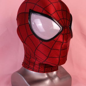 Pode incluir: Uma m&aacute;scara de tecido vermelho com um padr&atilde;o de teia de aranha preta na superf&iacute;cie. A m&aacute;scara inclui grandes lentes oculares brancas cobertas por uma malha e emolduradas por bordas pretas grossas. Exibida num busto de manequim prateado.