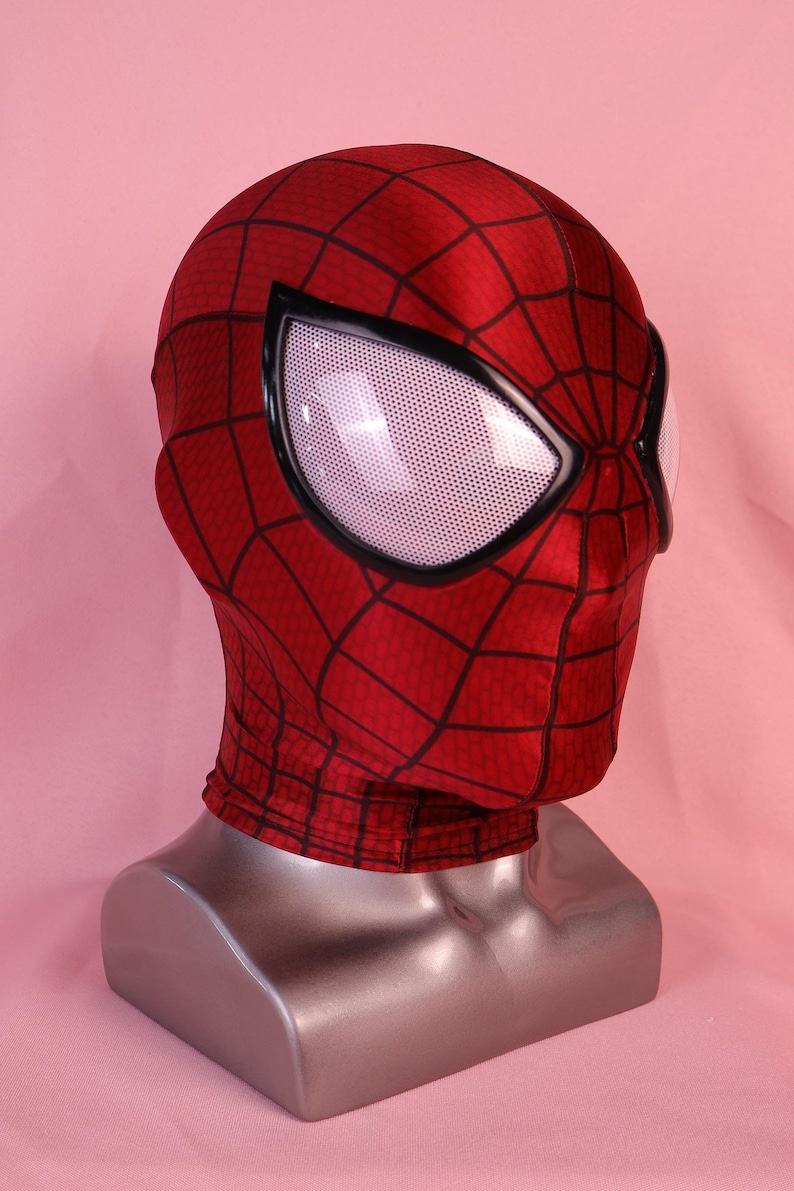 Pode incluir: Uma m&aacute;scara de tecido vermelho com um padr&atilde;o de teia de aranha preta na superf&iacute;cie. A m&aacute;scara inclui grandes lentes oculares brancas cobertas por uma malha e emolduradas por bordas pretas grossas. Exibida num busto de manequim prateado.