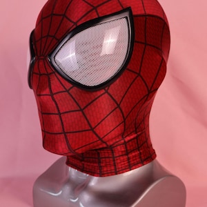Pode incluir: Uma m&aacute;scara de tecido vermelho com um padr&atilde;o de teia de aranha preta em toda a superf&iacute;cie. A m&aacute;scara inclui grandes lentes oculares angulares com uma textura de malha branca emolduradas por bordas pretas espessas.