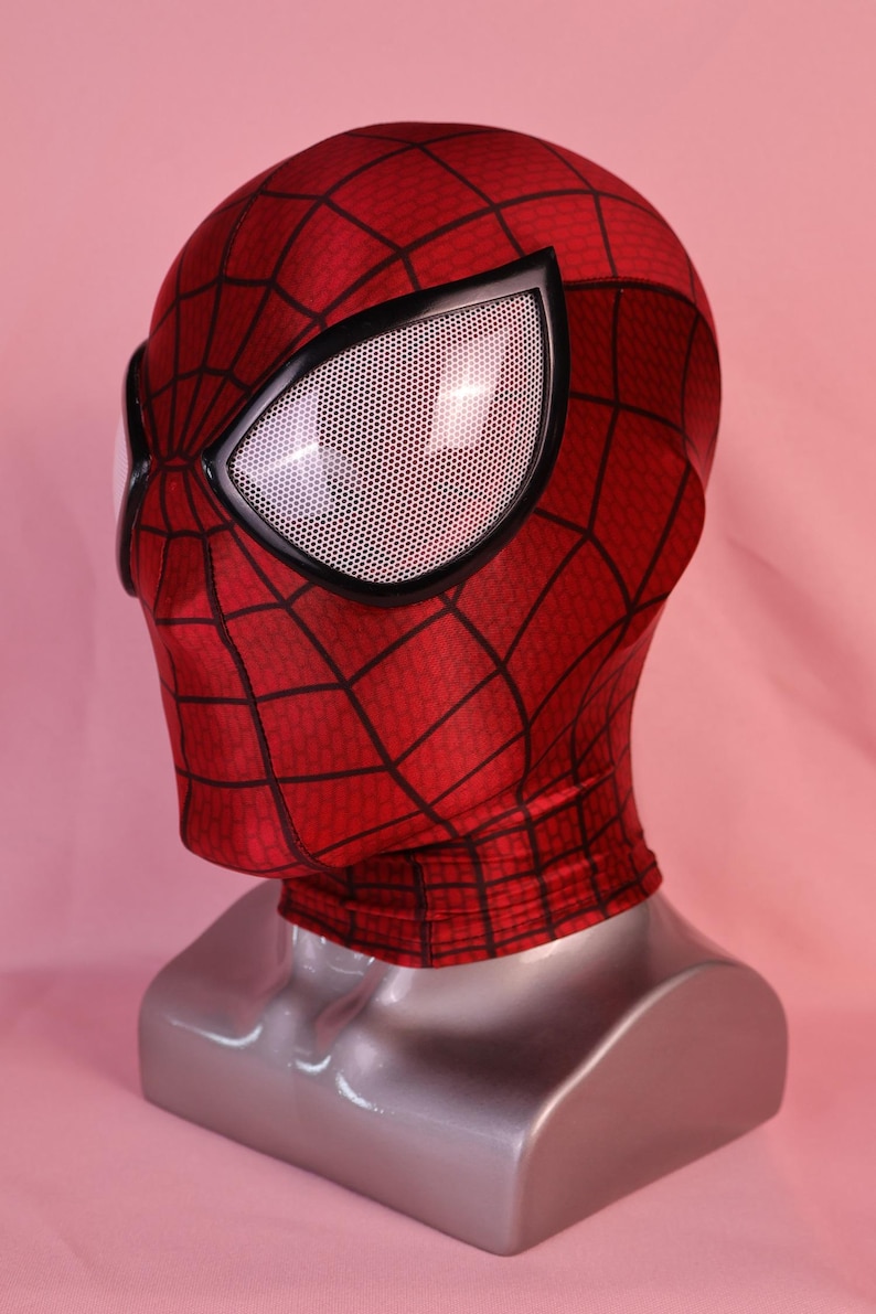 Pode incluir: Uma m&aacute;scara de tecido vermelho com um padr&atilde;o de teia de aranha preta em toda a superf&iacute;cie. A m&aacute;scara inclui grandes lentes oculares angulares com uma textura de malha branca emolduradas por bordas pretas espessas.