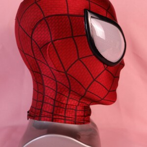 Pode incluir: Uma m&aacute;scara de tecido vermelho com um padr&atilde;o de teia de aranha preta em toda a superf&iacute;cie. A m&aacute;scara inclui lentes oculares grandes em forma de am&ecirc;ndoa com uma textura de malha branca e uma borda preta espessa, cobrindo cabe&ccedil;a e pesco&ccedil;o.