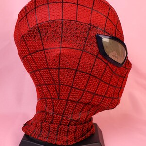 May include: A red full-head mask featuring a black webbed pattern across the surface. The mask includes a single visible eye lens with a metallic gold mesh texture framed in a dark blue border, designed to cover the head and neck area completely.