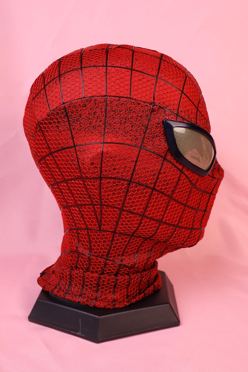 May include: A red full-head mask featuring a black webbed pattern across the surface. The mask includes a single visible eye lens with a metallic gold mesh texture framed in a dark blue border, designed to cover the head and neck area completely.