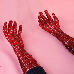 May include: A pair of long, red fabric gloves featuring a silver web pattern across the fingers, palms, and forearms. The gloves extend past the wrists and are constructed from a textured material with a distinct grid-like design throughout.