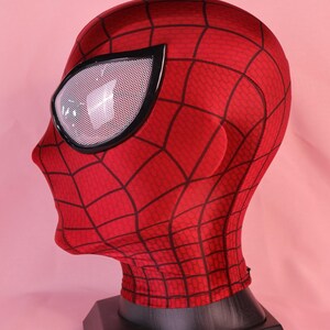 Pode incluir: Uma m&aacute;scara de tecido vermelho com um padr&atilde;o de teia de aranha preta na superf&iacute;cie. A m&aacute;scara inclui grandes lentes oculares brancas cobertas por malha e emolduradas em preto, cobrindo toda a cabe&ccedil;a e pesco&ccedil;o, apoiada num suporte preto.
