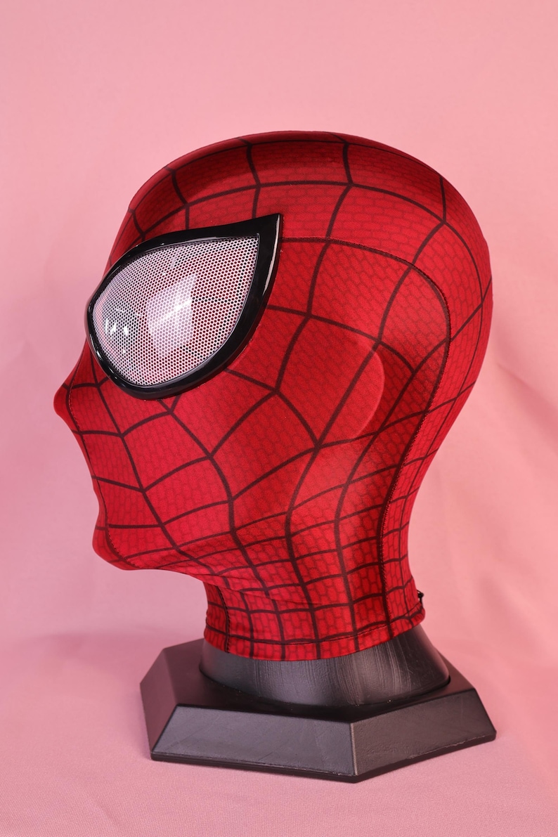 Pode incluir: Uma m&aacute;scara de tecido vermelho com um padr&atilde;o de teia de aranha preta na superf&iacute;cie. A m&aacute;scara inclui grandes lentes oculares brancas cobertas por malha e emolduradas em preto, cobrindo toda a cabe&ccedil;a e pesco&ccedil;o, apoiada num suporte preto.