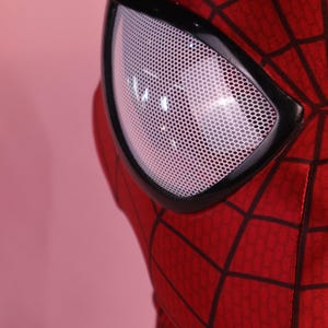 Pode incluir: Uma m&aacute;scara de tecido vermelho com um padr&atilde;o de teia preta na superf&iacute;cie. A m&aacute;scara inclui uma lente ocular triangular grande com uma textura de grade branca de malha fina, emoldurada por uma borda preta lisa que contorna o olho.