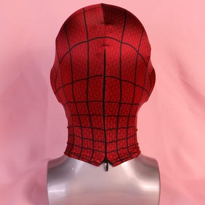 Pode incluir: Uma m&aacute;scara de tecido vermelho com um padr&atilde;o de teia de aranha preta impresso na superf&iacute;cie. A m&aacute;scara &eacute; exibida num manequim de cabe&ccedil;a e pesco&ccedil;o prateado, com um fecho de correr preto vertical ao longo do centro da parte traseira.