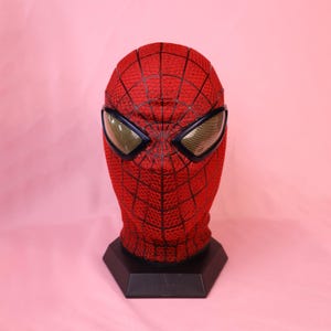 May include: A red full-head mask featuring a black web pattern across the surface. The mask includes two large, angular eye lenses with a gold-toned mesh texture, framed in black, mounted on a black hexagonal display stand for structural support.