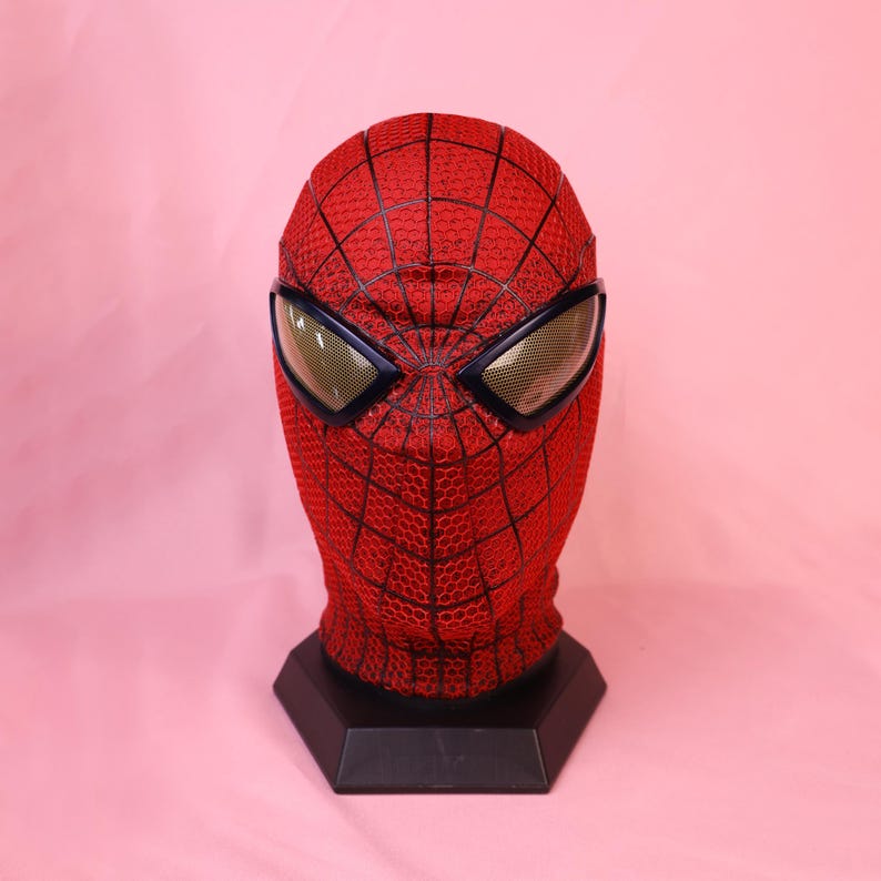 May include: A red full-head mask featuring a black web pattern across the surface. The mask includes two large, angular eye lenses with a gold-toned mesh texture, framed in black, mounted on a black hexagonal display stand for structural support.