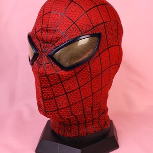 May include: A red fabric mask with a black web pattern and a textured honeycomb surface. The mask features large, angular eye lenses with a gold-toned mesh finish, mounted on a black hexagonal display stand for a full head-shaped presentation.