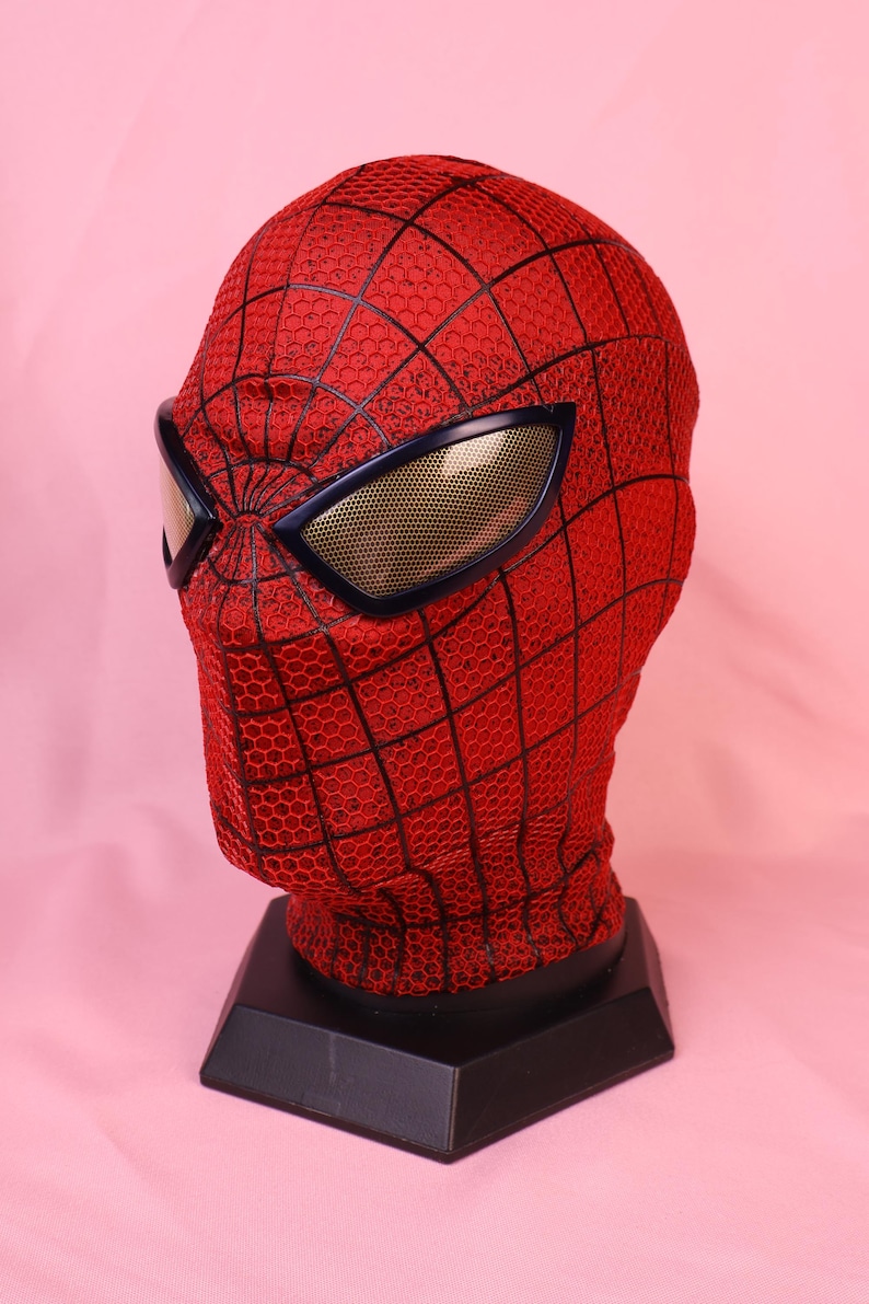 May include: A red fabric mask with a black web pattern and a textured honeycomb surface. The mask features large, angular eye lenses with a gold-toned mesh finish, mounted on a black hexagonal display stand for a full head-shaped presentation.