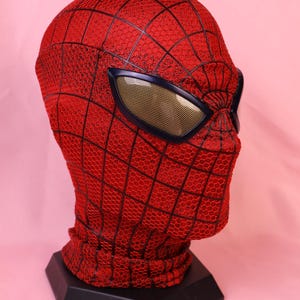 May include: A red full-head mask featuring a black webbed pattern across the surface. The mask includes large, angular eye lenses with a dark frame and a gold-toned mesh interior. The fabric has a textured, honeycomb-like appearance throughout.