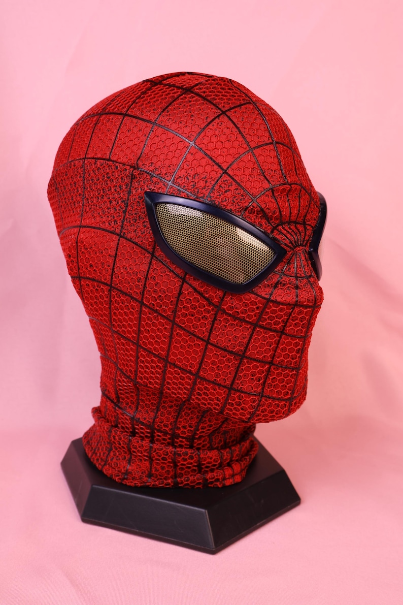 May include: A red full-head mask featuring a black webbed pattern across the surface. The mask includes large, angular eye lenses with a dark frame and a gold-toned mesh interior. The fabric has a textured, honeycomb-like appearance throughout.