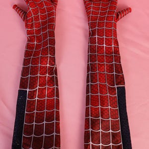May include: A pair of long, elbow-length gloves featuring a vibrant red fabric base with a printed silver web pattern. The gloves have a textured appearance and include a vertical black panel along the outer forearm of each piece.