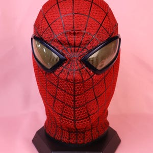 May include: A red full-head mask featuring a black webbed pattern across the surface. The mask includes two large, angular eye lenses with a gold-toned mesh texture framed in black, mounted on a black hexagonal display stand.