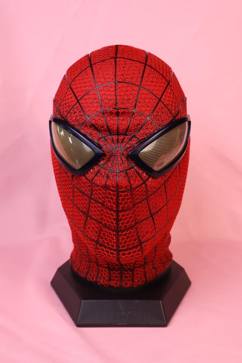 May include: A red full-head mask featuring a black webbed pattern across the surface. The mask includes two large, angular eye lenses with a gold-toned mesh texture framed in black, mounted on a black hexagonal display stand.