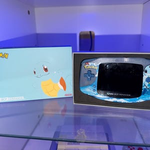 May include: A Game Boy Advance console and box set featuring the Pokémon character Squirtle. The box is light blue with the Pokémon logo and Squirtle illustration. The console has a Squirtle design and the words "Game Boy Advance".